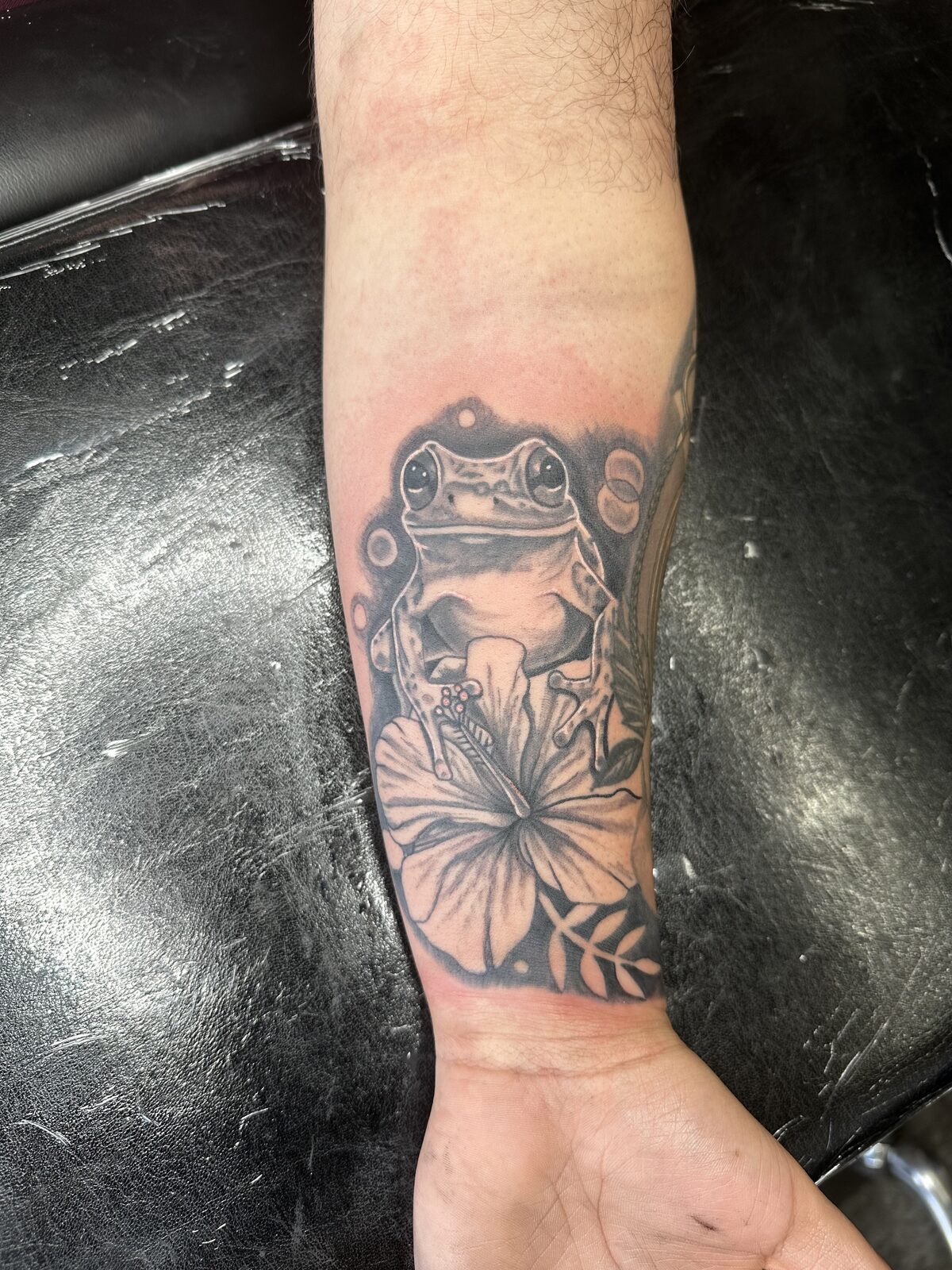 Black and gray frog sitting on a large flower with botanical leaf elements tattoo on an inner forearm